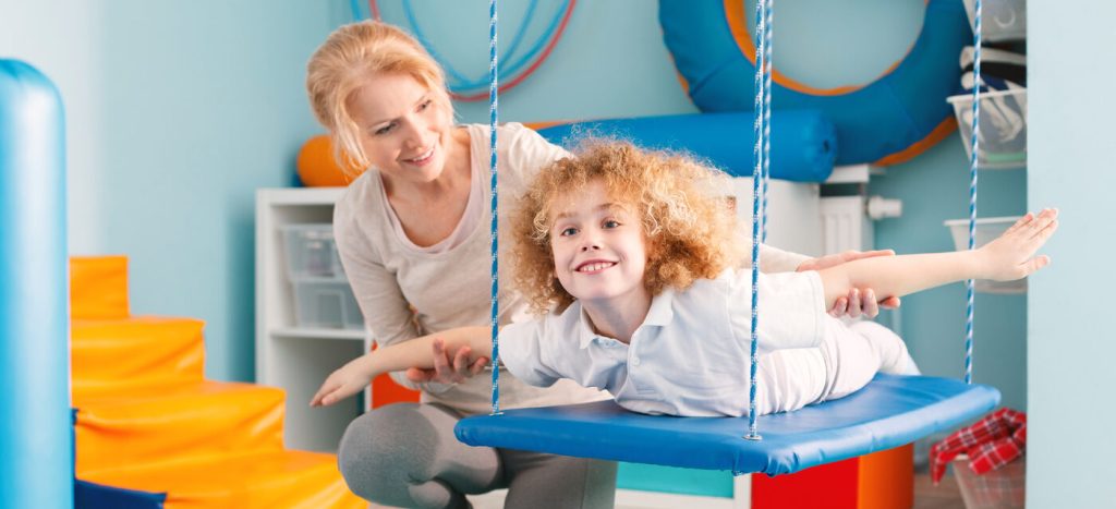 Woman helping a smiling boy to exercise on a therapy swing